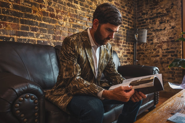 Man in a camouflage jacket reading a book on a leather couch against a brick wall.
